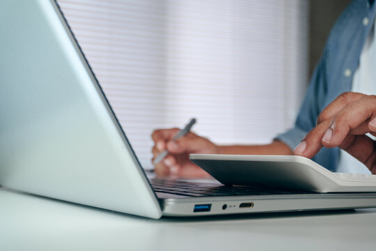 Close up of businessman hand using calculator on laptop computer and holding pen in modern office background, concept of business accounting, financial audit, and budget strategy.