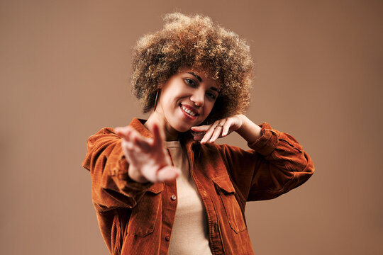 Smiling young woman with curly hair reaches toward the camera in a playful pose, wearing a rust jacket and beige top against a warm brown studio background.
