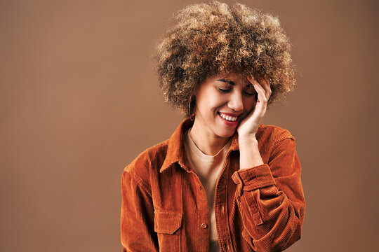 Smiling young woman with natural curly hair poses in an orange corduroy shirt against a warm brown background, touching her face in a relaxed studio portrait.