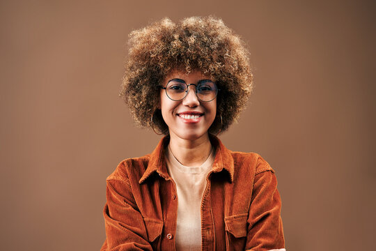 Smiling young woman with curly afro hair and round glasses wearing a rust corduroy shirt against a warm brown studio background.