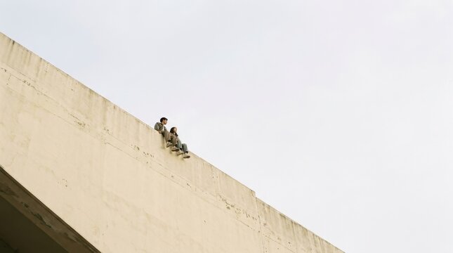 Two people sitting on the edge of a cream wall against pale sky, minimalist lifestyle photography. A couple perches at the top of a diagonal beige building, tiny against a vast pale gray-white sky,