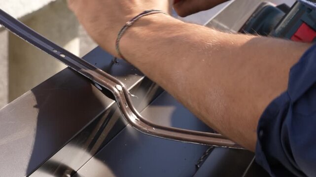 A builder installs a metal rain gutter on a roof using a drill and a rivet gun. A professional worker assembles and secures the components of a dark-brown metal gutter system on the exterior of a buil