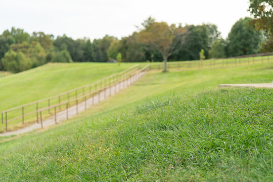 Shady side of a grass hill next to a slanted walkway in a public park with a paved path leading down from the parking lot to the recreation fields and beach area