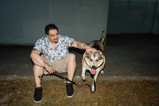 Man with tattooed arms sitting on curb petting dog at night