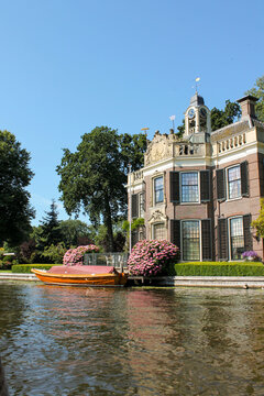 Exterior of a stately luxury villa with a large clock and sculptures on the facade, featuring a wooden sloop boat moored in front, a manicured hedge, and a garden with blooming pink rhododendrons