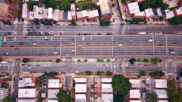 Top down aerial view of a multi lane highway passing over urban streets. Automobiles move along an elevated roadway above crosswalks, parking rows, and residential rooftops.