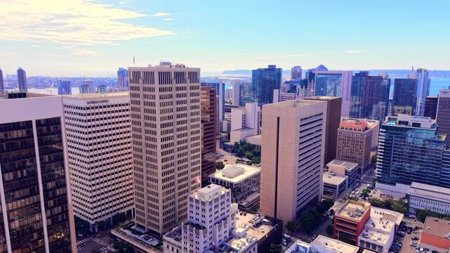 Office towers with American flags, downtown San Diego. Concrete and glass facades with rows of windows, rooftop antennas, tree lined streets and the bay on the horizon.