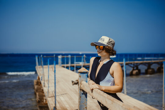 Young woman in cap and sunglasses posing on wooden pier by the sea during summer vacation