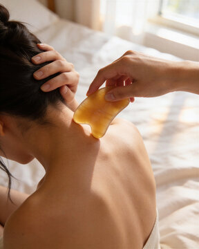 Woman experiencing gua sha scraping neck technique from therapist using yellow amber stone to relieve muscle tension in neck and back.