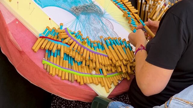 Artisan handcrafting intricate blue Valencian bobbin lace using wooden bobbins and pins on a lacemaking pillow during a traditional craft festival.