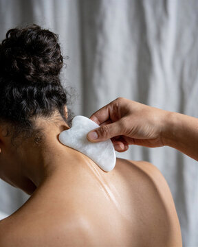 Professional therapist performing gua sha scraping neck technique with white jade tool on female client to relieve spine tension.