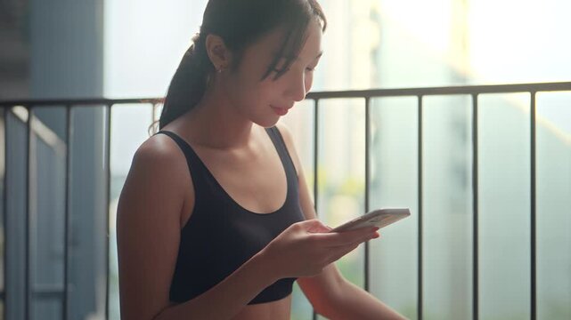 Close up of young Asian woman in black sportswear using smartphone while relaxing outdoors after exercise, female athlete checking mobile app in warm sunlight, fitness technology.