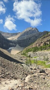 POV walking on rocky trail along Lake Agnes shore, then camera pans to emerald water below Big Beehive slope in Banff, Alberta. Concept of alpine hiking tourism and mountain freshwater conservation.