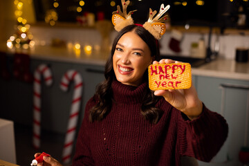 Happy woman wearing festive reindeer antlers decorating Christmas cookies and showing decorated biscuit to camera © Home-stock