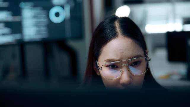 Data monitoring behind a foreground monitor, a woman in glasses watches multiple screens with blurred chart panels, her glasses catching light as the monitor edge frames the view across the office.