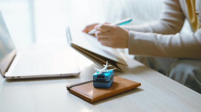 A person is writing on a tablet with a stylus beside an open laptop, with writing continuing behind a suitcase, model airplane and wallet that frame the travel planning concept at home.