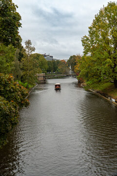 Small Boat on a Quiet City Canal Passing Under a Bridge Amid Autumn Trees and Overcast Sky