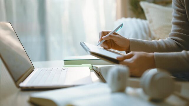 Digital notes take shape on a tablet as a hand uses a stylus for writing beside a laptop, with the tablet resting behind foreground books and headphones during a session of digital notes at home.