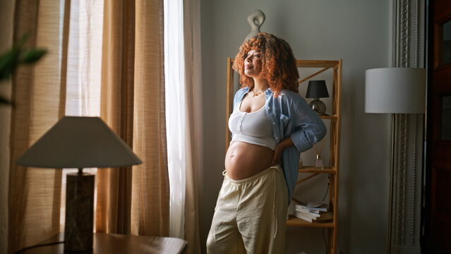 Smiling mom caressing belly window light home. Cozy maternity lifestyle indoors.