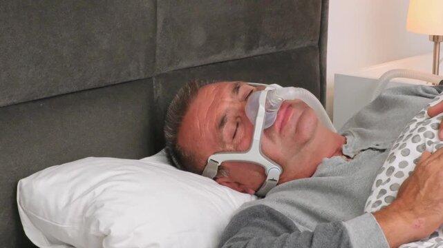 Senior Grey-Haired Man Sleeping in Bed Wearing a CPAP Mask, Sleep Apnea Device to Treat Obstructive Sleep Apnea and Prevent Breathing Pauses During the Night.