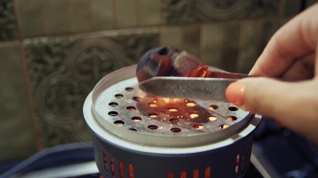 Person's hand turning a hot, red skull-shaped coal on a hookah burner with tongs