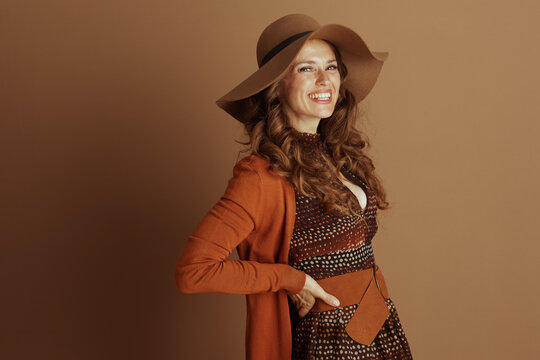A smiling woman in her 40s exudes confidence, posing with her hand on her hip in a chic autumnal ensemble. Her wide-brimmed hat, orange cardigan, and patterned dress reflect bohemian style.