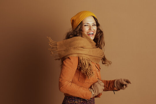 A joyful woman with curly brown hair, wearing a yellow beanie and orange jacket, smiles broadly and points forward against a neutral brown background, seen from the side.