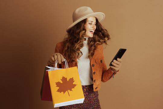 A joyful woman with curly brown hair, dressed in stylish autumn wear, laughs while looking at her smartphone and holding shopping bags with a prominent maple leaf design.