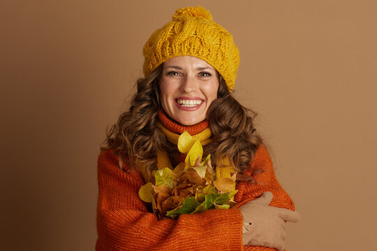 A cheerful woman with curly brown hair, dressed in warm orange and yellow autumn knits, smiles brightly at the camera while holding a bouquet of fall leaves.