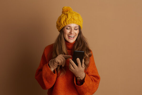 Cheerful woman in orange turtleneck and yellow pom-pom hat smiles, eyes closed, pointing joyfully at her smartphone. She embodies modern digital amusement and connection against warm brown background.