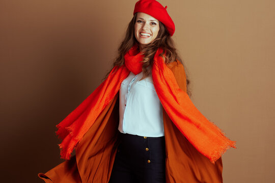 A vibrant woman wearing beret, scarf, and long orange-brown coat over blue pants, smiles joyfully with outstretched arms. Posed dynamically against warm background, she embodies stylish autumn cheer