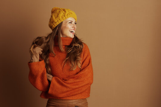 Joyful woman, long wavy brown hair, yellow beanie, orange turtleneck, brown pants, smiles and laughs, casually holding hair. Plain light brown background, embodying cheerful autumn fashion.