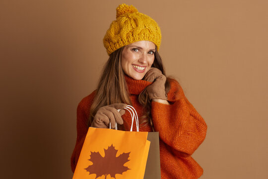 A smiling woman in her 30s, dressed in a yellow pompom hat and orange turtleneck, holds two autumn-themed shopping bags. Soft studio lighting enhances the warm, fashionable seasonal atmosphere.