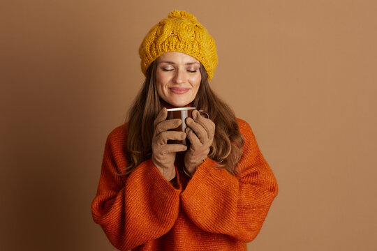 A woman with long brown hair, wearing a yellow knitted hat, orange sweater, and gloves, gently smiles with closed eyes while holding a dark mug. The scene evokes a cozy, peaceful autumn atmosphere.