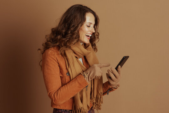 A smiling woman with long curly brown hair, wearing a warm orange jacket and brown scarf, points to her smartphone screen, engaged in modern communication.