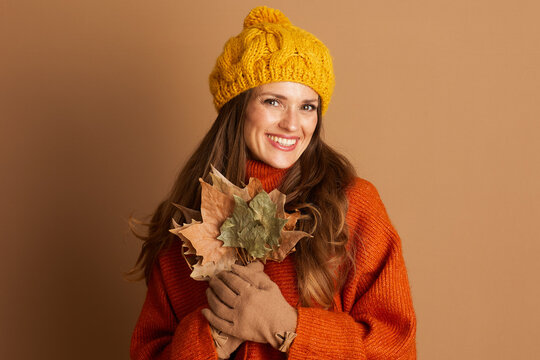 A joyful woman in a yellow beanie and orange sweater holds autumn leaves, looking directly at the camera. This warm portrait captures the cheerful spirit of the season.