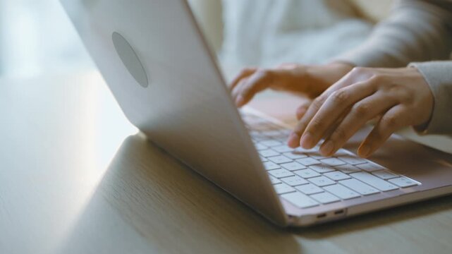 Two hands continue laptop typing on a keyboard beside a softly lit sofa, with fingers pressing keys in a side view as laptop typing and home computing create a quiet domestic device routine.