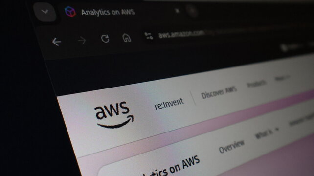 AWS website homepage presented on computer monitor screen closeup view in dark room environment, showcasing cloud computing platform brand logo and top menu bar.