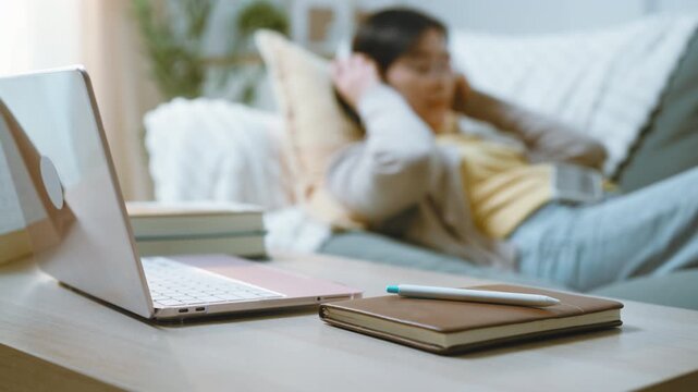 A woman is relaxing on a sofa while using a smartphone for mobile browsing, with an idle laptop, headphones and notebook nearby as she scrolls on the smartphone during a quiet moment at home.