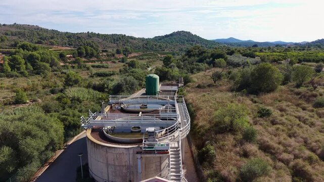 Compact municipal sewage treatment plant with oval aeration tank and circular clarifiers amid green hills and orchards, seen from above