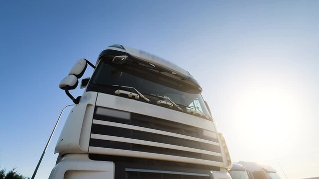 White Semi Truck Cab Parked in Row with Other Trucks at Truck Stop | Close-Up of Front Grille and Windshield | Ultra Wide Low Angle View, Blue Sky | Freight Transport Logistics Trucking