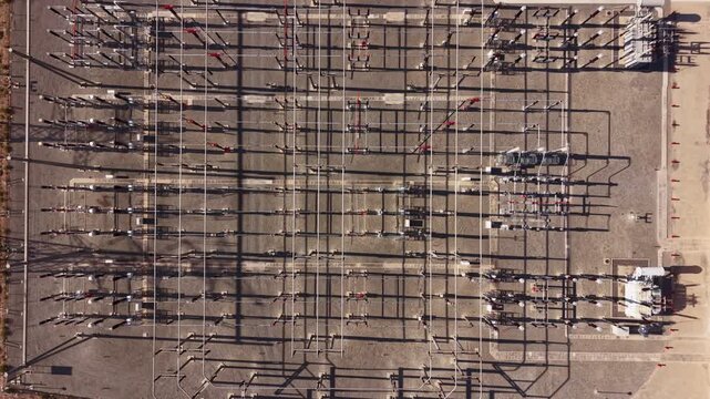 Overhead view of an electrical substation with rows of high-voltage equipment, switchgear and transmission structures casting long shadows on gravel ground