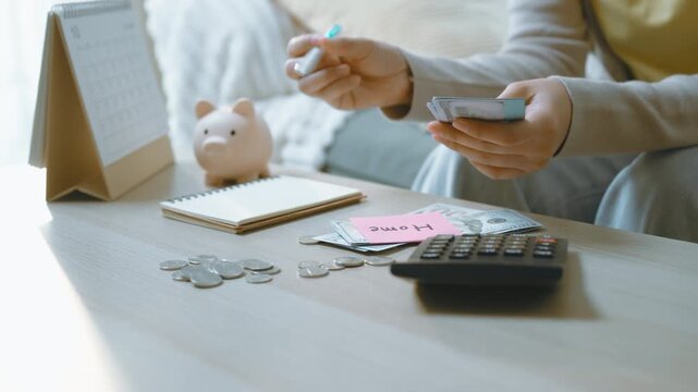Hands count banknotes while writing in a notebook beside coins, a calculator and piggy bank, with writing accompanying cash allocation as visible home and car labels establish separate savings goals.