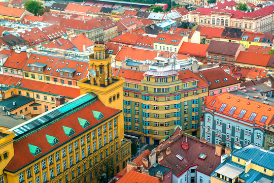 Elevated view across Prague's residential rooftops, showcasing historic architecture, orange tiles, pastel facades and surrounding green hills.