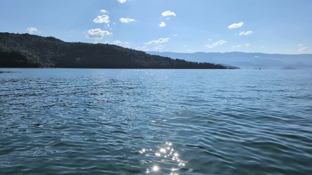 Handheld slow motion of calm blue sea water with sun glittering on the surface in Paraty, Rio de Janeiro, featuring lush tropical mountains under a sunny blue sky with soft white clouds.