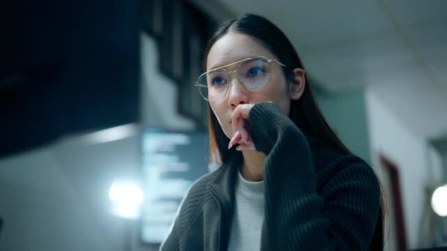 Yawning at work in a dim office, a woman in glasses holds a hand over her mouth near the monitor, her glasses catching light as the sustained yawning gives way to lowered eyes during a brief pause.