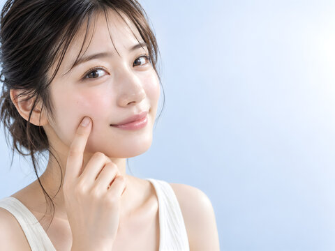 澄んだ瞳と明るい美肌が印象的な女性が頬の柔らかさを指先で確かめて微笑むスキンケア美容写真 淡いブルー背景に広がる右側のコピースペース