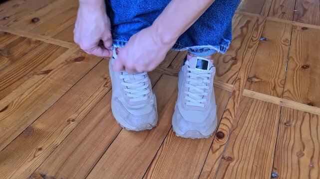 Person tying the laces of gray sneakers while standing on a wooden floor indoors