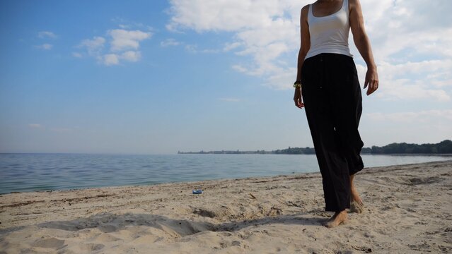 Happy adult woman going on sandy beach near shoreline at sunny day. Beautiful lady walking on seashore with calm waves at background. Concept of summer vacation or holiday. Slow motion