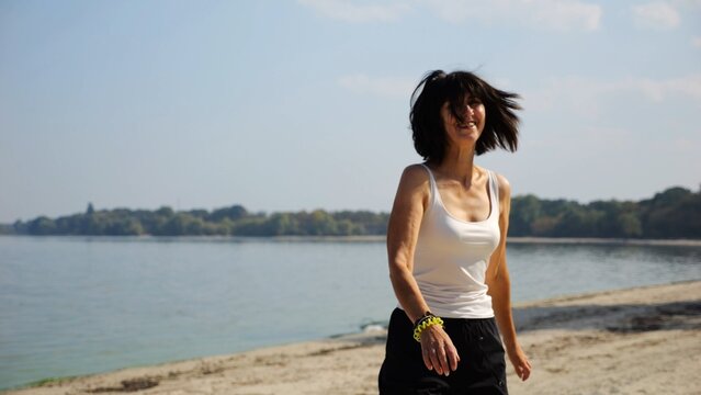 Beautiful lady walking on sandy beach near shoreline at sunny day. Happy adult woman turning around near coastline. Carefree female enjoying rest at seaside. Concept of summer vacation or holiday